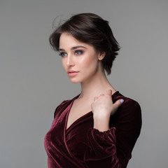 Fototapeta premium Beauty female portrait. Young attractive woman posing at studio over gray background. Beautiful model with perfect make-up dressed evening burgundy dress looking at camera.