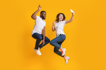 Fototapeta na wymiar Cheerful Interracial Couple Taking Selfie While Jumping In Air