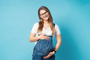 Fototapeta na wymiar Smiling pregnant woman wearing glasses caressing her belly over blue background