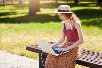 Obraz premium Outdoor shot of girl looking for information via laptop and wireless Internet, charges with help of solar panel built in wooden bench, looks concentrated, doing online work. Alternative energy concept