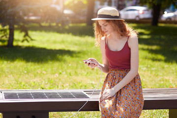 Young beautiful woman having rest in park, sitting on innovative bench and charging mobile device while checking her social network, using solar panel, Ecology and modern technology concept.