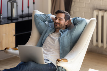 Peaceful young man daydreaming in comfortable armchair, holding computer.