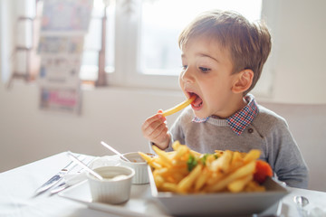 Portrait of small little cute caucasian boy kid eating french fries potato chips at the table in the restaurant or at home three or four years old