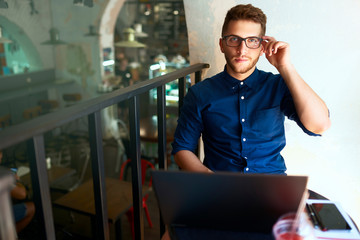 Authentic portrait of young confident businessman looking at camera with laptop in office. Hipster man in glasses doing his startup project in modern loft open workspace. Business telecommuting theme.