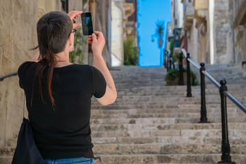 Naklejka premium Traveler woman taking photo with mobile phone of Medieval narrow street with stairs in Valletta, Malta.