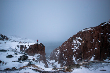 frozen waterfall on the background of hills on the Kola Peninsula and a man standing on the edge of the cliff and looking into the distance to the Arctic ocean