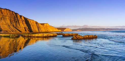 Sunset view of the Pacific Ocean shoreline, with golden colored cliffs reflected on the wet sand, Drakes Beach, Point Reyes National Seashore, California