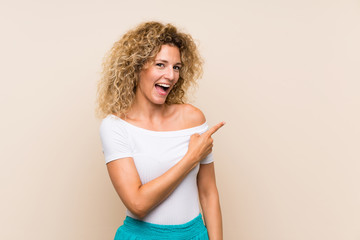 Young blonde woman with curly hair over isolated background surprised and pointing side