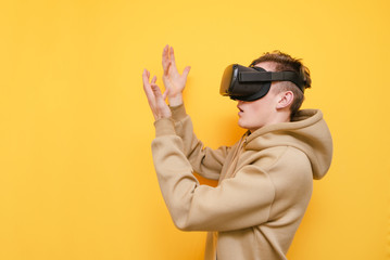 Portrait of surprised guy in VR helmet stands on yellow background and looks at his hands. Young...