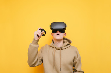 Portrait of surprised young man in VR helmet stands on yellow background with controller in hand and looks up in amazement. Guy gamer plays video games on VR helmet. Isolated.