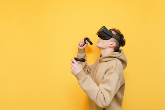 Portrait of a concentrated young man with a VR helmet on his head and controllers in his hands on a yellow background, playing video games in virtual reality and look away at copy space