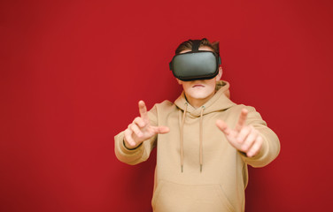 Serious guy in VR helmet stands on a red background with a smile on his face and points his hands at the camera. Young man wants to touch a picture in a helmet of virtual reality.