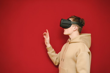 Handsome man in casual clothes stands on a red background with a VR helmet on his head, looks away and touches his finger to copy space. Teenager uses VR technology, wears a virtual reality helmet.