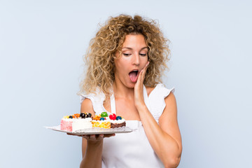 Young blonde woman with curly hair holding lots of different mini cakes with surprise and shocked facial expression