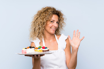 Young blonde woman with curly hair holding lots of different mini cakes saluting with hand with happy expression