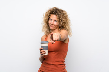 Young blonde woman with curly hair holding a take away coffee points finger at you with a confident expression
