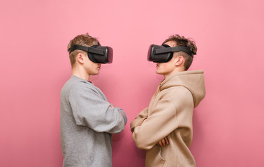 Two young men in casual clothes stand facing each other in VR helmets on their heads and look at...