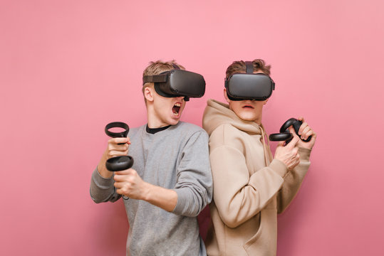 Two emotional young men in VR helmets compete in a virtual reality game. Two gamers in virtual reality helmets play VR games, hold controllers. VR gaming, leisure with friends