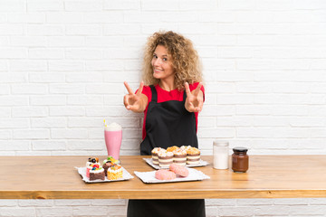 Young woman with lots of different mini cakes in a table smiling and showing victory sign