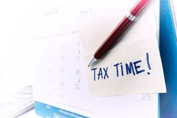 Tax time concept; hand is holding Tax Time written on the white paper note with a red pen and calendar.