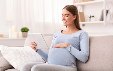 Pregnant Lady Using Tablet Reading E-Book Sitting On Sofa Indoor