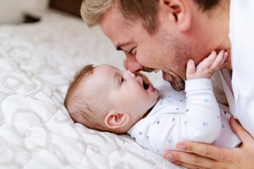 Close up of cute six months old baby boy lying on bed and playing with his dad. Dad talking to son...