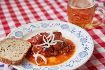 Czech food specialty, roasted small sausages in beer sauce made from dark beer, tomato sauce, chily, garlic and onion served on rustic plate with glass of lager beer and bread, typical czech pub snack