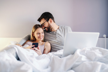 Young attractive couple sitting on bed in bedroom and cuddling. Man having laptop in lap while woman holding smart phone.