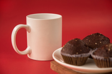 A white Cup of coffee stands on a wooden Board. Chocolate cupcakes are on the plate. Red background.
