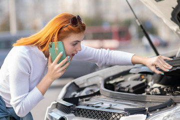 Young woman standing near broken car with popped hood talking on her mobile phone while waiting for help.