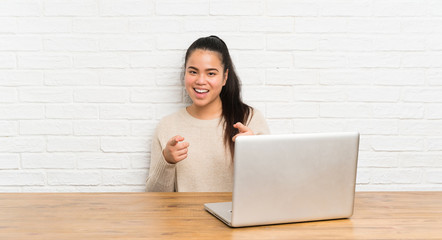 Young teenager Asian girl with a laptop in a table points finger at you