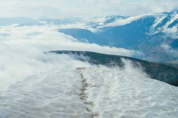 Footprints in snow on mountain top above thick clouds with view to giant mountains and glaciers. Snow on mountain peak among thick low clouds. Atmospheric alpine landscape. Wonderful highland scenery.