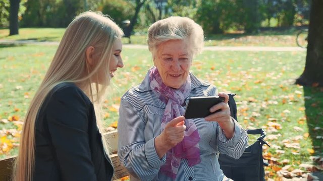 Beautiful blonde girl with elder woman sitting on bench and looking something on smartphone. They spending time in a big autumn park with golden leaves on a grass.
