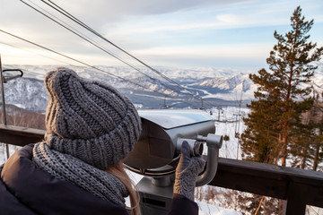 Obraz premium A young girl in a gray knitted hat looks through stationary binoculars on an observation deck in the alatai mountains in winter with snow.