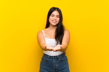 Young teenager Asian girl over isolated yellow background holding copyspace imaginary on the palm...