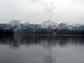 Obraz premium Cloudy winter day in the city. Houses, trees and bridges in the city are reflected in the water of the river on a cloudy day at the beginning of winter.