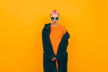 Extraordinary beautiful woman with short pink hair in dark coat and sunglasses smiles isolated on orange background