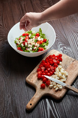 Red сherry tomatoes with salad cheese on cutting board. Cooking vegetable salad. Female hand, knife, food ingredients and a white ceramic bowl