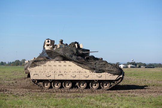 Fighting vehicle used by US army is moving on field and battlefield. Machine has camouflage over turret. Unrecognizable soldier is on the top. Pannign shot with slightly blurred background.