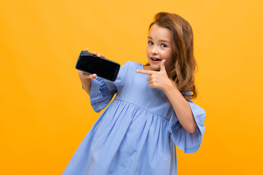 Cute Smiling Girl In A Blue Dress Shows The Phone In Horizontal Position With A Mockup On A Yellow Background With Copy Space