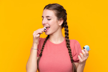 Young woman over isolated yellow background holding colorful French macarons and eating it