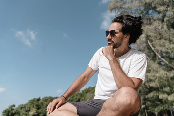Naklejka na ściany i meble Handsome sexy latin brunette man with a beard in sunglasses and a white T-shirt in profile against a tropical landscape