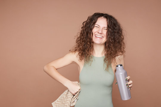 Carefree positive European lady dressed in active wear, holds bottle of fresh water, glad to be healthy, has regular training, carries soft towel in hand, isolated on beige studio wall with copy space