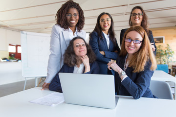 Coworkers using laptop and smiling at camera. Group of cheerful multiethnic female colleagues working with laptop computer in office. Women in business concept