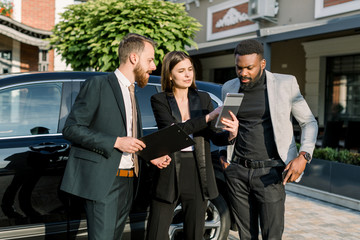Two men, African and Caucasian, and one Caucasian woman stand in front of black car on a parking place on yard of dealer. Woman showing something on the digital tablet