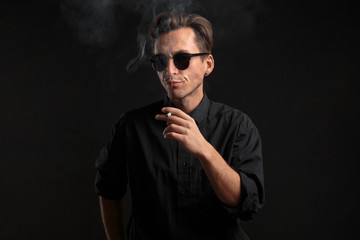 Stylish young man in a black t-shirt and sun glasses smoking a cigarette over black background.