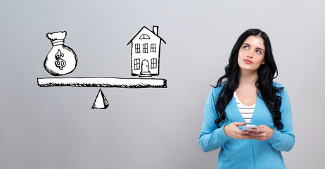 House and money on the scale with thoughtful young woman holding a smartphone