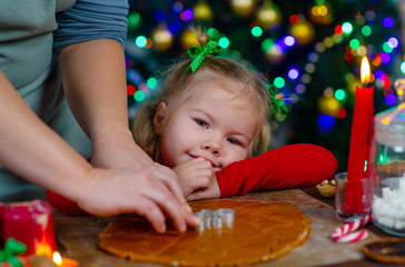 Little girl watches as mom cuts cookies out of dough with a cookie cutter, against the background of a Christmas tree
