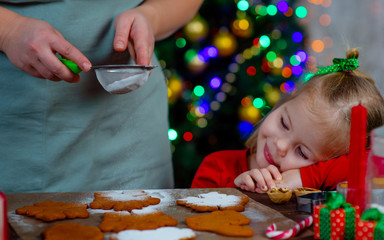 A little girl watches as mom sprinkles powdered cookies on a Christmas tree background
