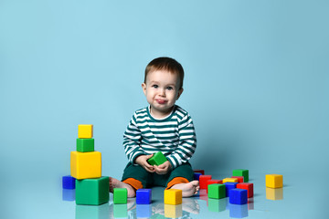 Obraz premium Little baby boy in stylish casual clothing barefoot sitting on floor, playing with colorful toy cubes, showing tongue